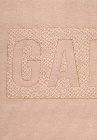 Tessuto beige con lettere in rilievo e cucite che formano la scritta "GAP" su uno sfondo testurizzato. Le fibre mostrano una finitura liscia e uniforme.