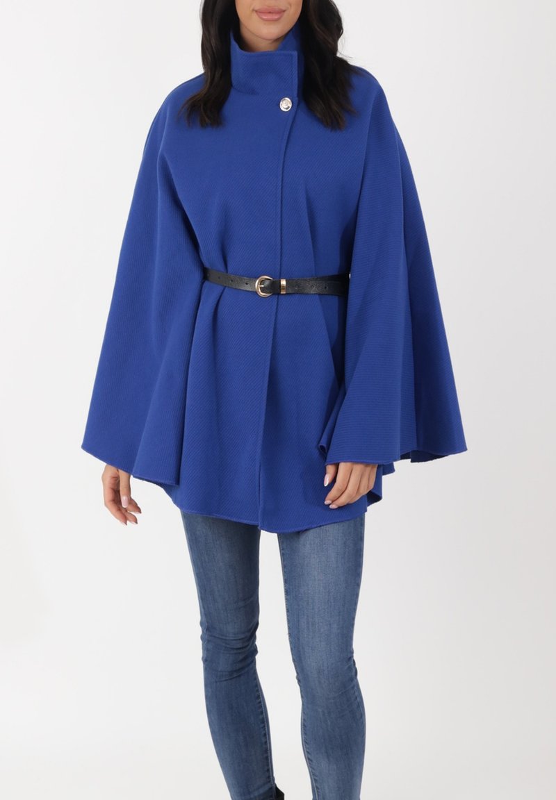 Blå cape-kappa med struktur, hög krage, en silverknapp och svart bälte i midjan, bärs över blå jeans.