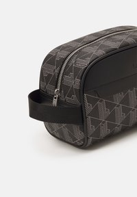 Svart toalettmappe med glidelås, geometrisk preget mønster og stoffhåndtak. Har glatte sider og en matt finish på toppen.