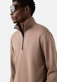 Příliš světle hnědý pulovr s krátkým zipem, vyrobený z hladké látky s vysokým límcem a kontrastním zipem. Nositelný s reflexními slunečními brýlemi.