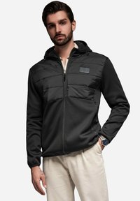 Giacca nera con zip, corpo in maglia testurizzata e pannello imbottito sul petto. Presenta un cappuccio e una toppa con logo sulla parte superiore sinistra. Assenza di motivi.