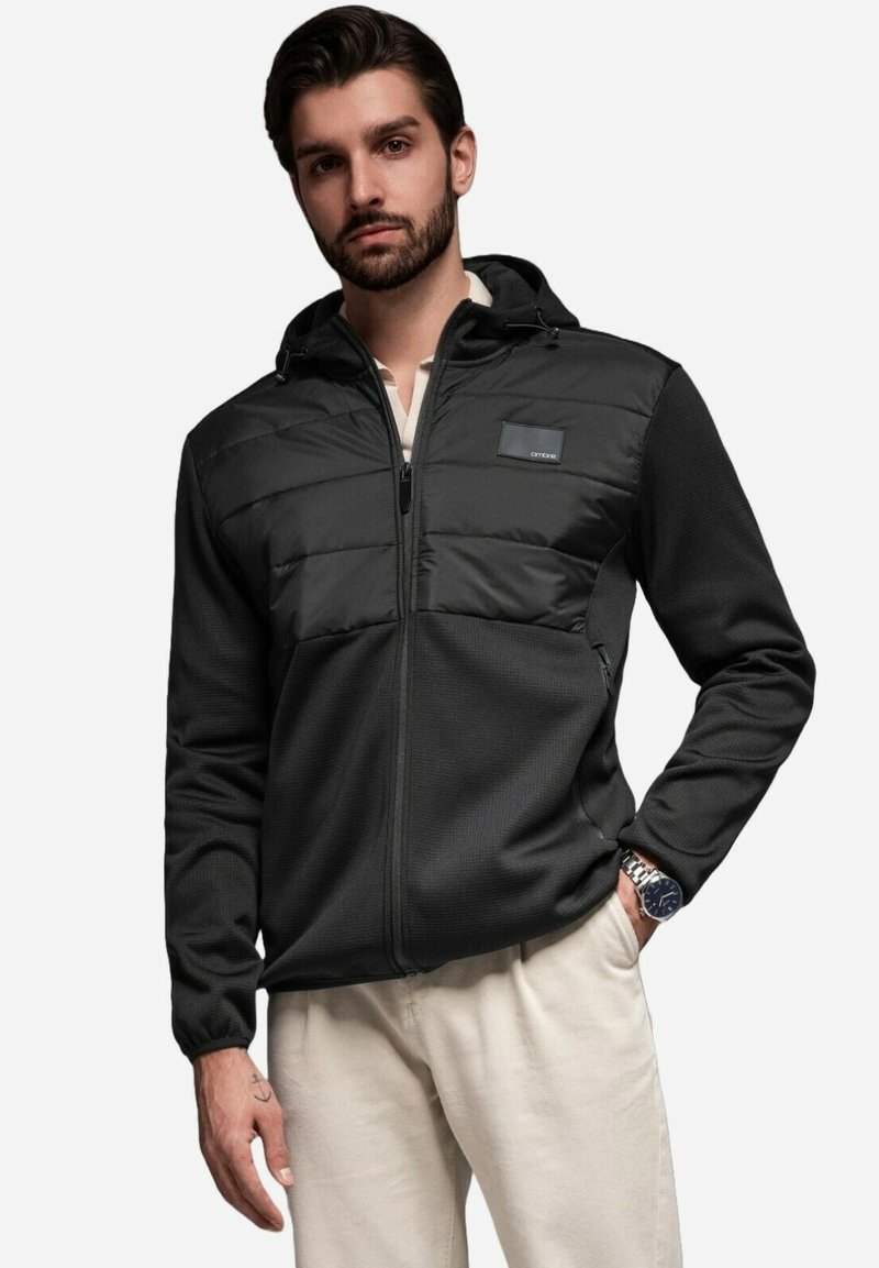 Giacca nera con zip, corpo in maglia testurizzata e pannello imbottito sul petto. Presenta un cappuccio e una toppa con logo sulla parte superiore sinistra. Assenza di motivi.