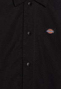 Juodas marškinis su mygtukais, pagamintas iš tekstūruotos medžiagos, turi smailų apykaklę, kontrastingą siuvinėjimą ir mažą Dickies logotipo etiketę ant krūtinės.