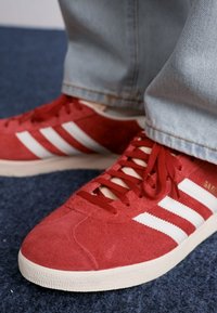 Sarkani zamšas sporta apavi ar baltām svītrām, aprīkoti ar plakaniem sarkaniem aukliņām, teksturētu virsu un gumijas zoli. Akcentēti ar zeltainu zīmolu.