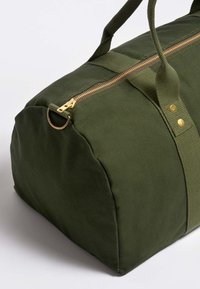 Grøn canvas gymtaske med en gylden lynlås, rund form, stofhåndtag og messinghardware detaljer. Glat tekstur med forstærkede sømme.