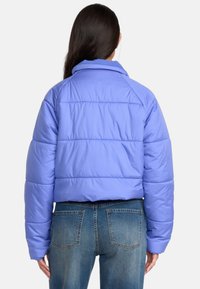 Geknipte pufferjack in licht lavendel met horizontale stiksels, brede kraag en lange mouwen. Gecombineerd met hooggetailleerde blauwe jeans.