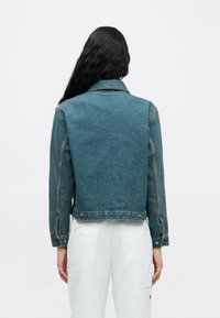 Άτομο με μακριά σκούρα μαλλιά, φοράει μπλε τζιν μπουφάν και λευκό παντελόνι, στέκεται γυρισμένο με την πλάτη προς το απλό λευκό φόντο.