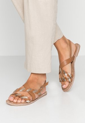 Fødder i beige bukser iført brune åbne sandaler med fletede metalliske og læder remme samt ankelspænder på en hvid gulv.