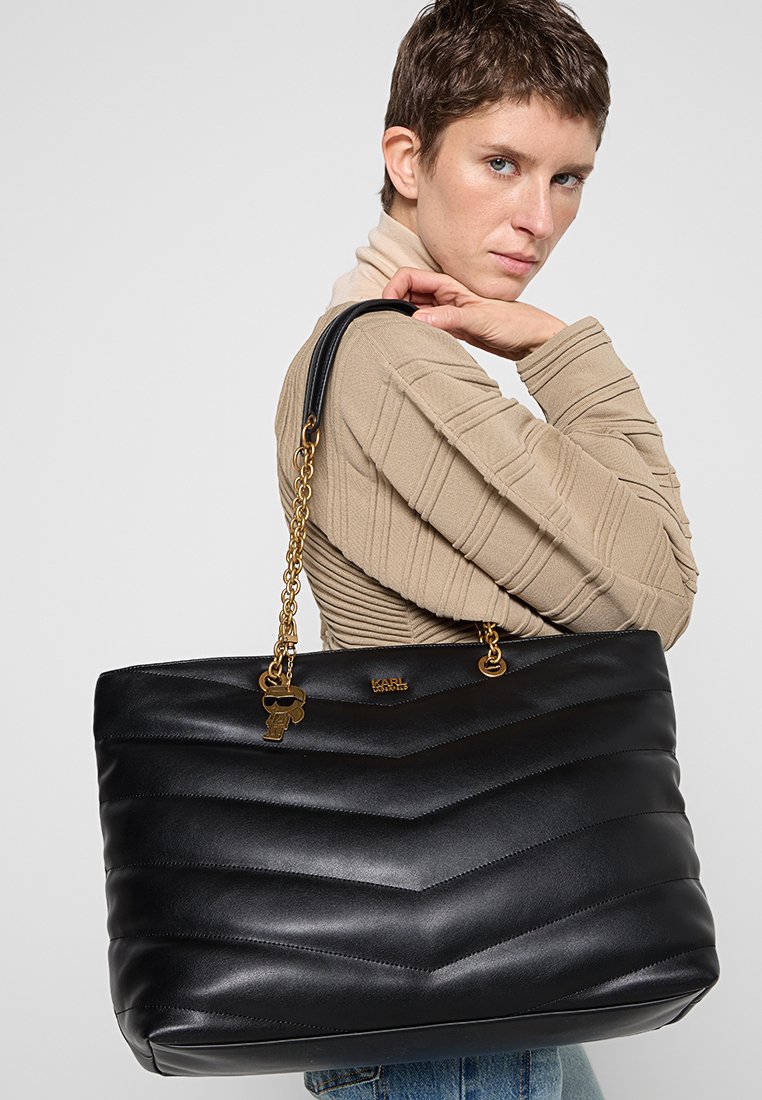 Bolso tote de cuero negro con patrón acolchado en zigzag, correa de cadena dorada y adorno de llavero. Presenta una forma estructurada y un interior espacioso.