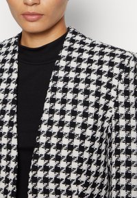 Svart och vit houndstooth-mönstrad blazer i texturerat tyg, med notchade slag och öppen framdesign.