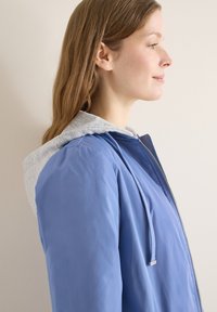 Jeune femme de profil portant une veste bleue avec une capuche grise, debout devant un mur uni de couleur claire.