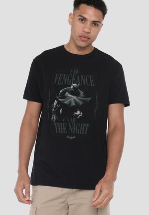 Junger Mann trägt ein schwarzes Batman-T-Shirt mit dem Text ""I am vengeance, I am the night"" und einer Grafik von Batman mit ausgebreitetem Umhang.