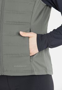 Grå vadderad väst med dragkedja framtill och sidofickor, med en slät yta och "ENDURANCE"-logotyp vid fållen.