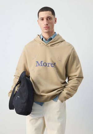 Joven vestido con sudadera con capucha beige con texto "More!", camisa azul claro debajo, pantalones crema, sosteniendo una bolsa negra contra un fondo liso.