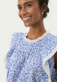Blusa a fantasia floreale blu con maniche a volant e raffinato pizzo bianco. Tessuto leggero con una consistenza morbida e dettagli arricciati.