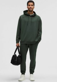 Tummanvihreä huppari, jossa on huppu ja narut, yhdistettynä yhteensopiviin jogger-housuihin. Malli pitää mustaa laukkua. Valkoiset lenkkarit viimeistelevät asun.