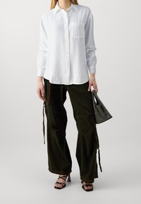 Chemise blanche à manches longues avec boutons, en tissu texturé ; portée avec un pantalon cargo ample vert foncé et tenant un petit sac noir en mesh.