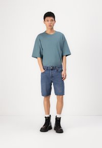 Camiseta de algodón oversized en color azul verdoso con mangas cortas, combinada con pantalones cortos de mezclilla azul y botas negras con cordones por el tobillo. Atuendo simple y casual.