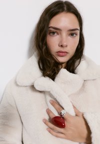 Krém színű műszőrme kabát nagy gallérral, egy ezüst gyűrűt viselő kéz és egy fényes piros, szív alakú kiegészítő díszíti.