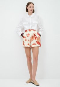 Witte langemouwen shirt met voorzakken, bloemenpatroon shorts in crème en rode tinten, en beige instap schoenen. Geen zichtbare accessoires.