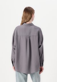 Camisa oversized de botão em tecido cinza suave com finas riscas verticais e um detalhe de laço nas costas. Mangas longas com punhos abotoados.
