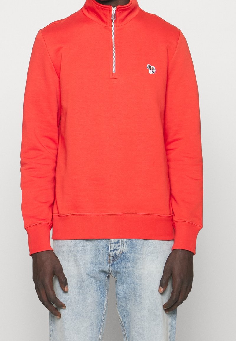 Sweat-shirt rouge zippé avec col montant, manches longues et poignets côtelés. Présente un petit logo sur la poitrine. Associé à un jean bleu clair.