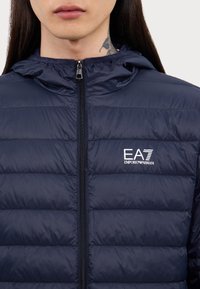 Veste rembourrée marine avec fermeture éclair à l'avant et capuche. Présente des coutures horizontales, un logo sur le côté gauche et une texture lisse et brillante.