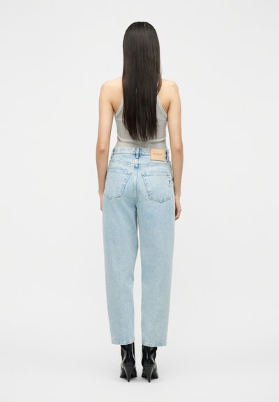 Γυναίκα με μακριά ίσια μαλλιά που φοράει γκρι αμάνικη μπλούζα, ανοιχτόχρωμα τζιν με ψηλή μέση και μαύρα κοντά μποτάκια με τακούνι, φαίνεται από πίσω.