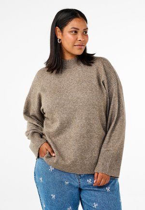 Brauner, flauschiger Strickpullover mit hohem Kragen, überschnittenen Schultern und gerippten Bündchen, kombiniert mit blauen Jeans mit weißen Schleifenmustern.