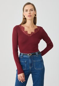 Burgunderfarget langermet topp med ribbet tekstur og V-hals med blonder. Kombinert med høyhalsede denimjeans og et svart belte.