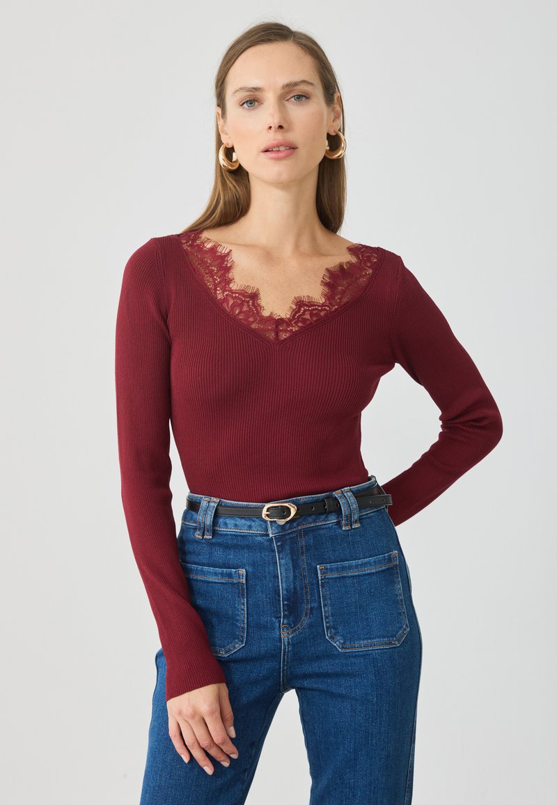 Burgunderfarget langermet topp med ribbet tekstur og V-hals med blonder. Kombinert med høyhalsede denimjeans og et svart belte.