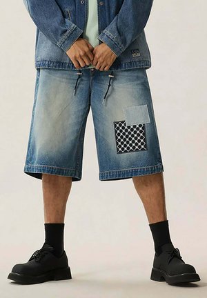Uomo che indossa pantaloncini di jeans sbiaditi con due toppe di stoffa, calzini neri, scarpe nere con lacci e una giacca di jeans abbinata.