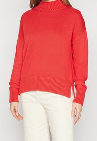 Rood gebreide trui met een hoge hals, geribbelde textuur, schuine zijsplitten en lange mouwen. Gecombineerd met lichtgekleurde broek.