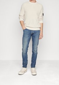 Ljust beige stickad tröja med ribbade manschetter och rund halsringning, matchad med åtsittande blåjeans och off-white skor med ljusa accenter.