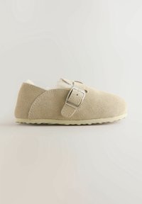 Chaussure beige en daim à enfiler avec une boucle argentée et une doublure blanche douce et duveteuse, vue de côté sur un fond neutre.