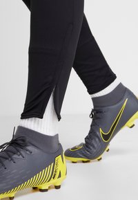 Zwarte sportleggingen met een ritsdetail bij de enkels, gecombineerd met grijze en gele Nike voetbalschoenen, met een subtiel grippatroon en veters.