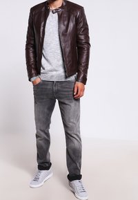 Brun läderjacka, grå tröja, grå jeans och vita sneakers. Jackan har en rund krage och dragkedjor på sidorna.