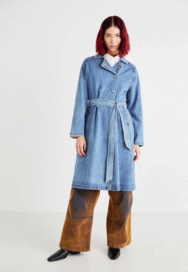 Trench coat in denim con vita regolabile, bottoni doppi, e un lavaggio azzurro chiaro; indossato sopra pantaloni larghi marroni.
