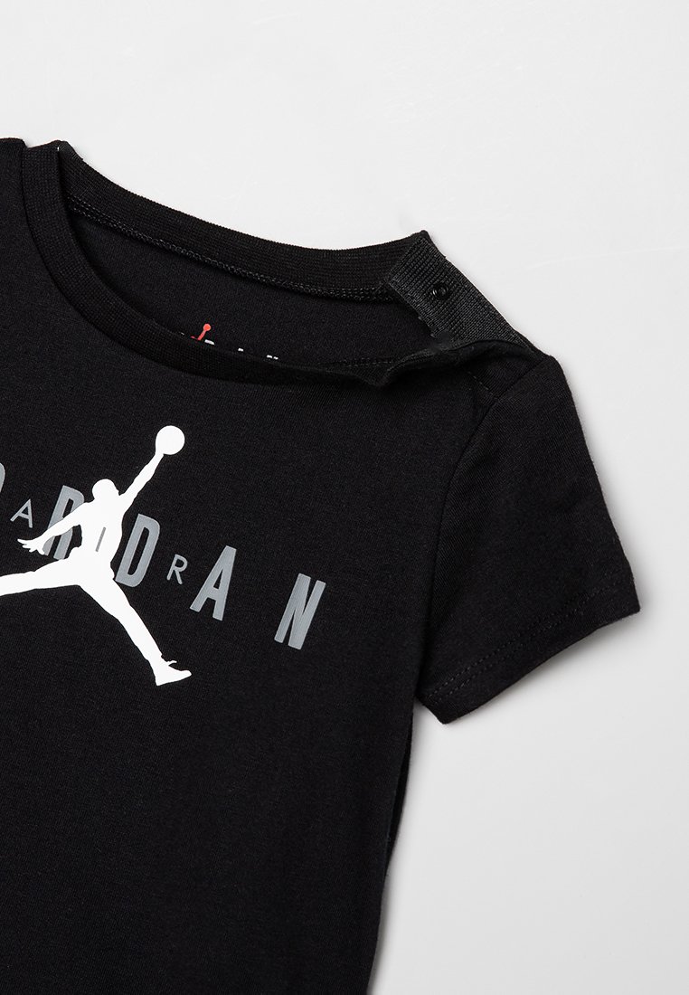 T-shirt en coton noir avec un logo Jumpman blanc et le texte "JORDAN" en gris. Manches courtes avec une fermeture à bouton-pression au niveau du col.