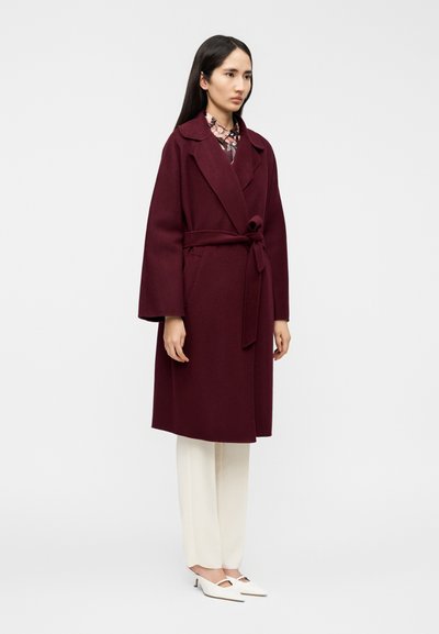 Μπουφάν από μαλλί σε μπορντό χρώμα με ζώνη, λαιμόκοψη με σχισμές και φαρδιά μανίκια, συνδυασμένο με κρεμ παντελόνια και λευκά παπούτσια με λεπτομέρειες κοψιμάτων.