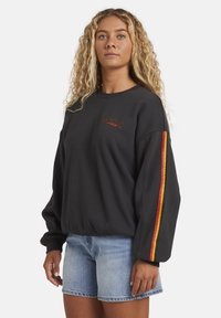 Sudadera negra de manga larga, con un pequeño logo bordado en el pecho y acentos a rayas coloridas a lo largo de las mangas.