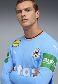 Hellblaues Sporttrikot mit langen Ärmeln, das das deutsche Nationalemblem, Logo-Patches und einen runden Halsausschnitt mit mehrfarbigem Rand zeigt.