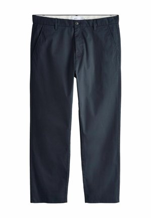 Mann trägt dunkelblaue Hose und weißes Hemd, mit rechter Hand entspannt an der Seite vor einfarbigem Hintergrund.