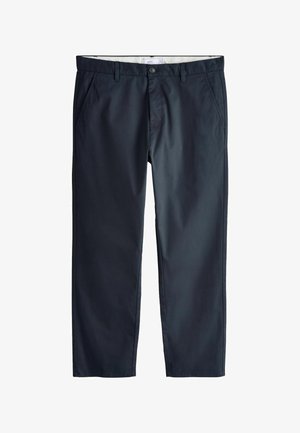 Mann trägt dunkelblaue Hose und weißes Hemd, mit rechter Hand entspannt an der Seite vor einfarbigem Hintergrund.