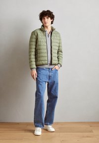 Giubbotto imbottito verde oliva con zip frontale, abbinato a una maglietta a maniche lunghe grigia, jeans blu larghi e sneakers bianche.
