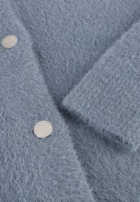 Lichtblauwe fuzzy cardigan met grote zilveren knopen en gebreide textuur. De stof is zacht en heeft een losse, casual uitstraling.