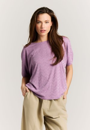 Vrouw met lang bruin haar, gekleed in een paarse top met korte mouwen en zigzagpatroon en beige broek, met handen in de zakken.