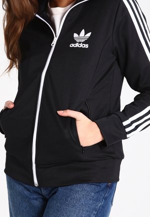 Veste de survêtement noire Adidas zippée avec des bandes blanches sur les manches et un logo, portée avec les mains dans les poches avant et un jean bleu.