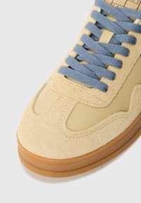 Zapatilla de ante y cuero beige con suela de goma, que presenta cordones de color azul claro y elementos de diseño curvados. Superficie texturizada en la zona de los dedos.