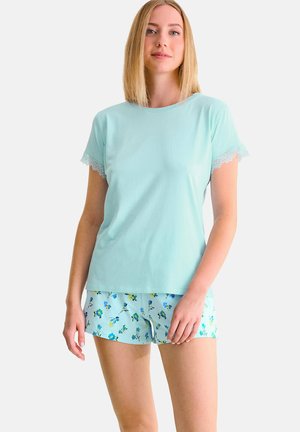 Femme portant un haut à manches courtes bleu clair avec bordure en dentelle et un short assorti à motif floral, debout devant un fond blanc uni.